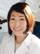 医療白衣を着た女性が微笑んでいるポートレート。背景には医学関連の資料が見える。