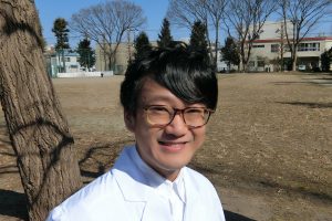 医療白衣を着た男性が公園で微笑んでいる様子。背景には木々と建物が見える。