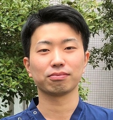 医療関係の服を着た男性が、緑の植物の前で微笑んでいる写真。