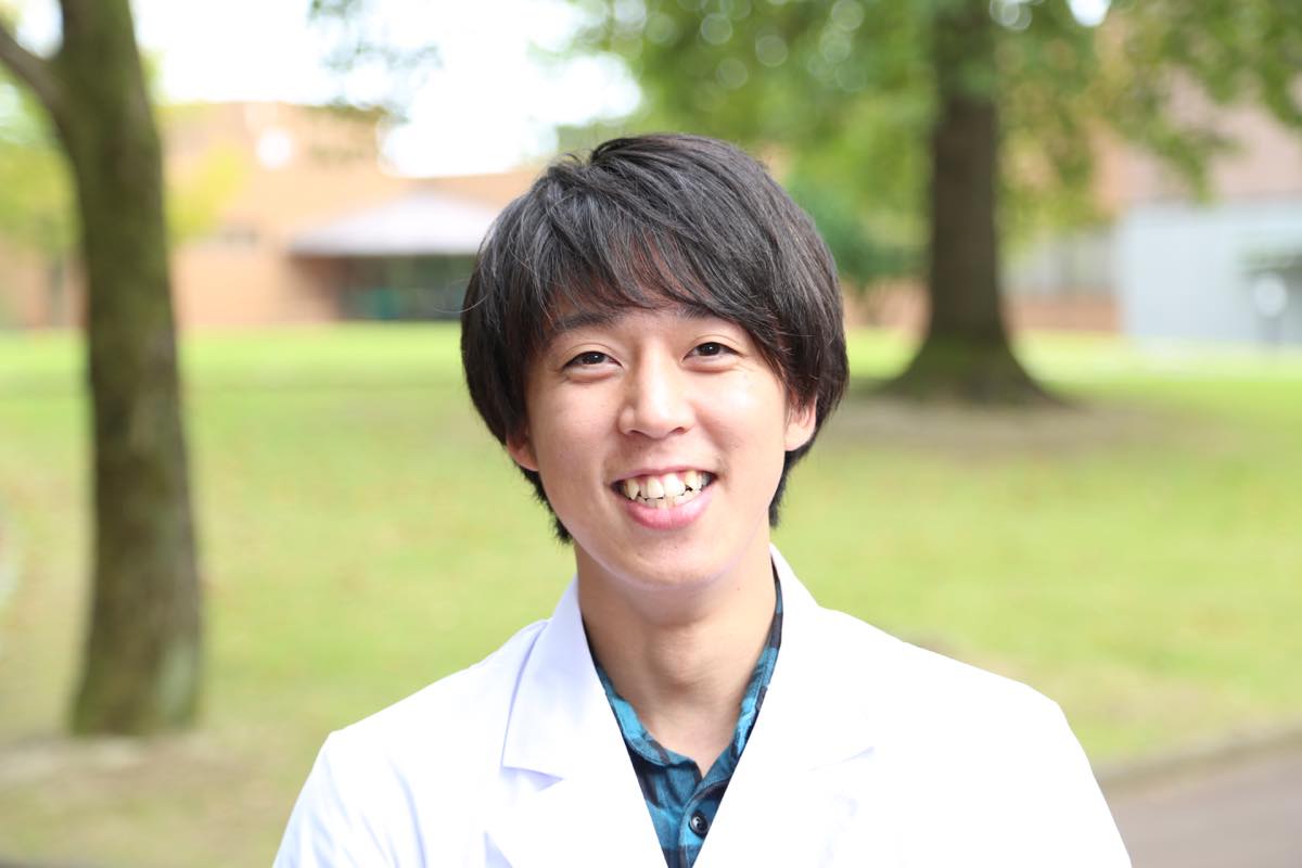 福島県立医科大学の白衣を着た男性が笑顔で立っている風景。背景には緑の木々が見える。