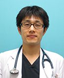 医療用白衣を着た男性が青い背景の前で微笑んでいる写真。