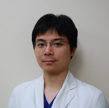 医療用白衣を着た男性がカメラに向かって微笑んでいるポートレート。背景は淡い色合いの壁。