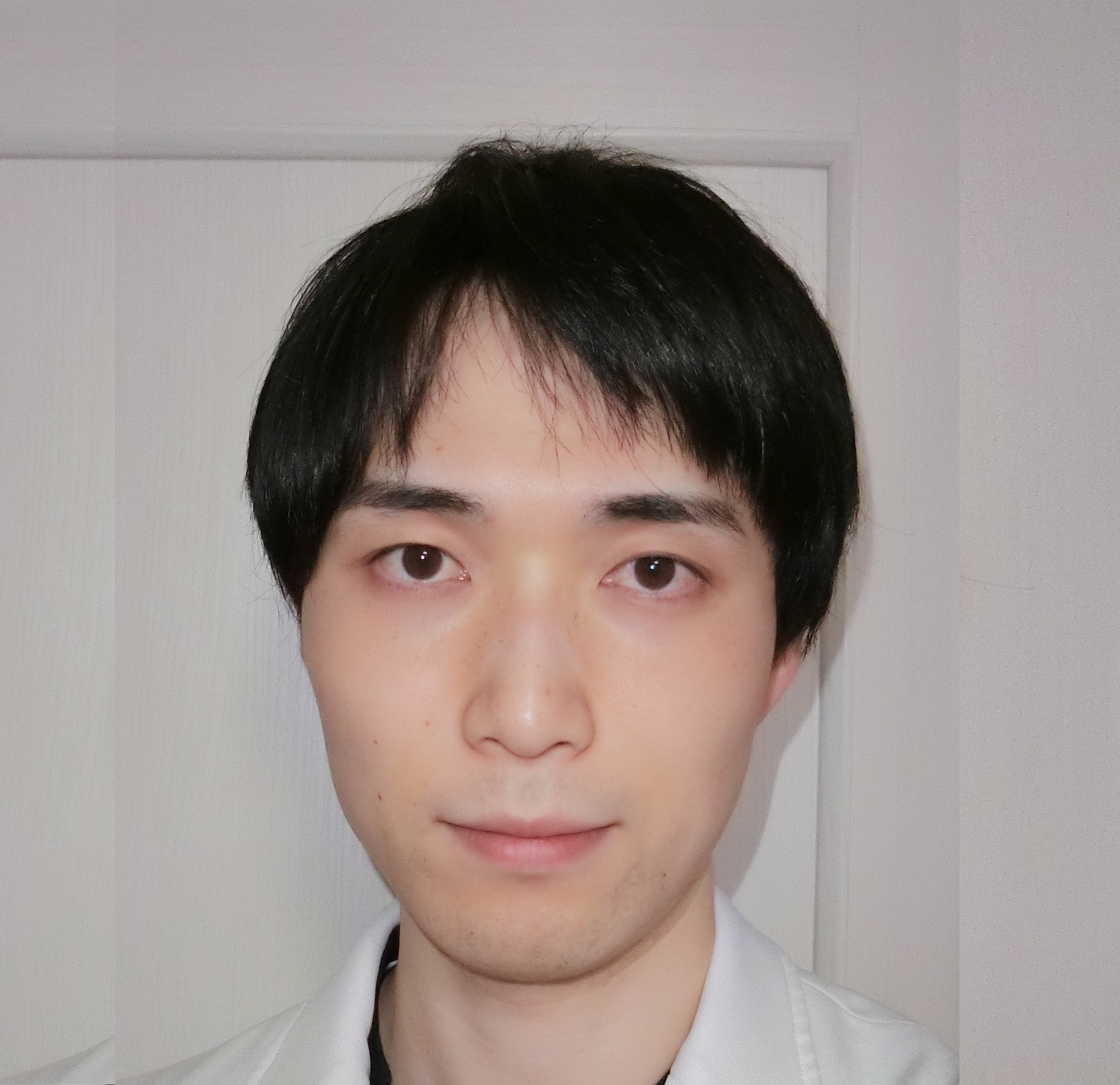 医療用白衣を着た若い男性の顔写真。背景は白い壁。