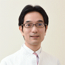 医療白衣を着た男性がカメラに向かって微笑んでいるポートレート。背景は淡い色合い。