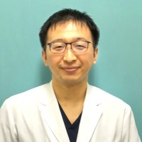 医療白衣を着た男性が青い背景の前で微笑んでいる写真。