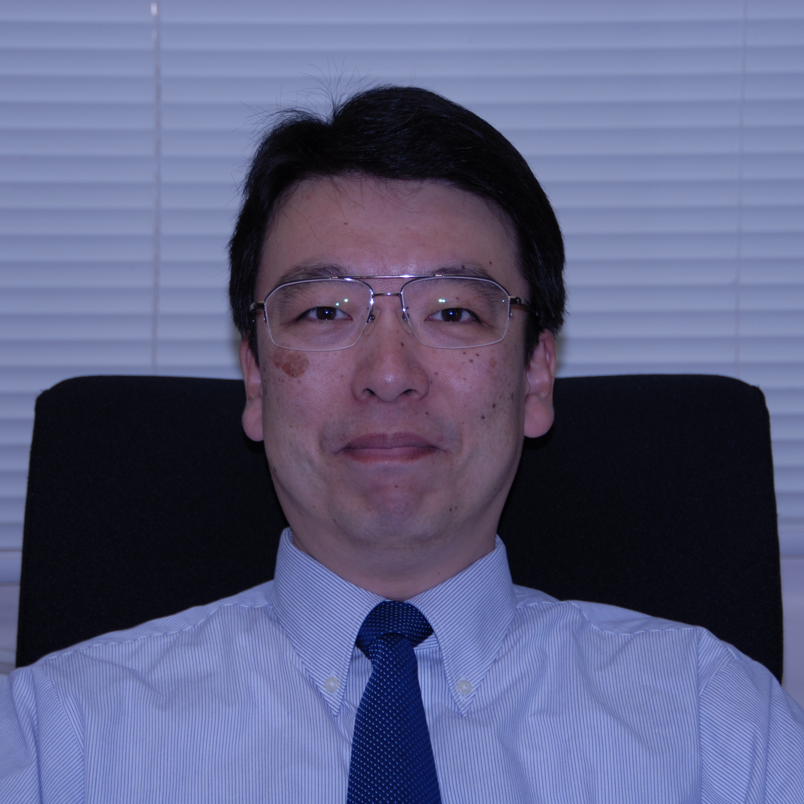 医療に従事する男性が椅子に座り、カメラに向かって微笑んでいるポートレート。背景にはブラインドが見える。