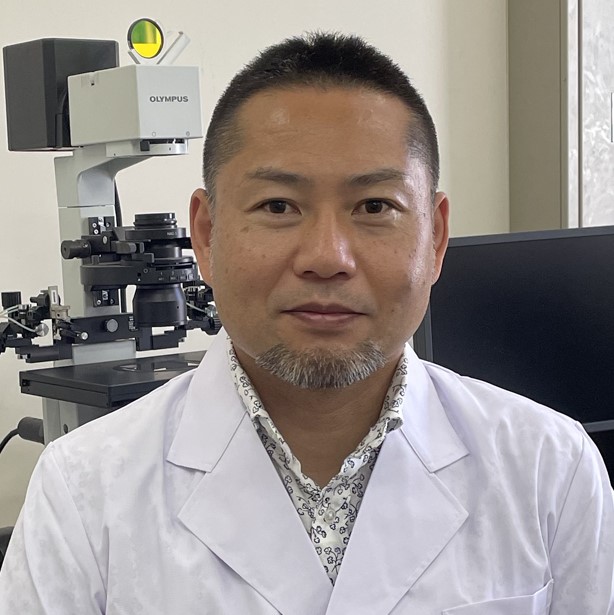 医学部の准教授が白衣を着て微笑んでいる写真。背景には顕微鏡とコンピュータが見える。