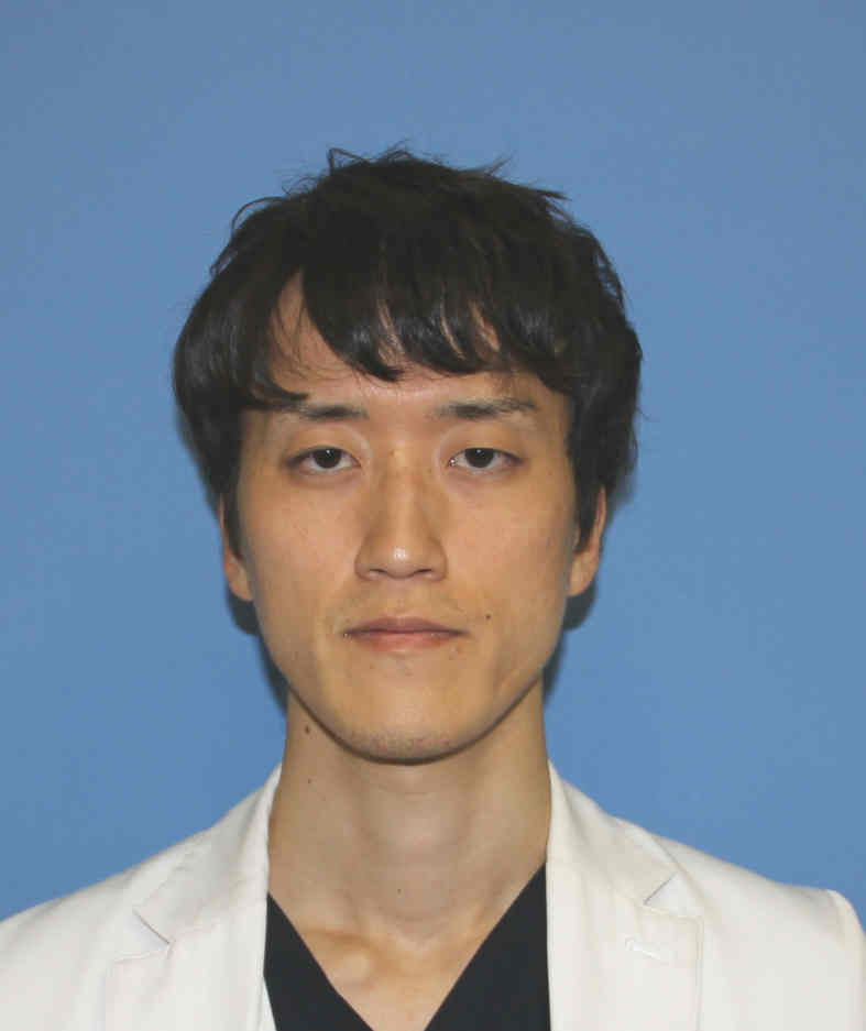 福島県立医科大学の松本拓朗助教のポートレート写真。白いジャケットを着用し、青い背景の前で真剣な表情を浮かべている。
