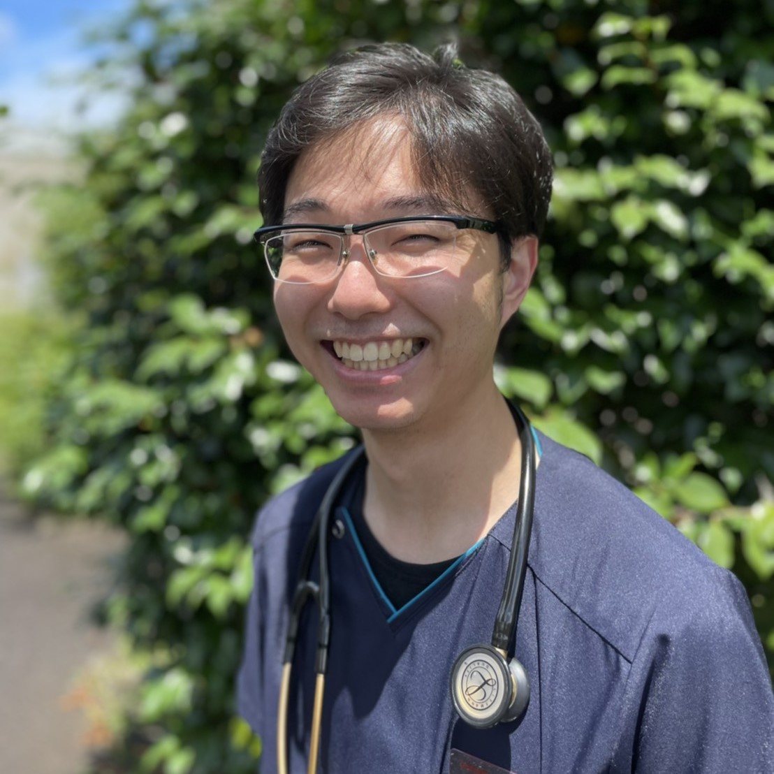 医療用の聴診器をつけた若い男性が、緑豊かな背景の前で笑顔を見せている。