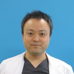 医療用白衣を着た男性が青い背景の前で微笑んでいる写真。