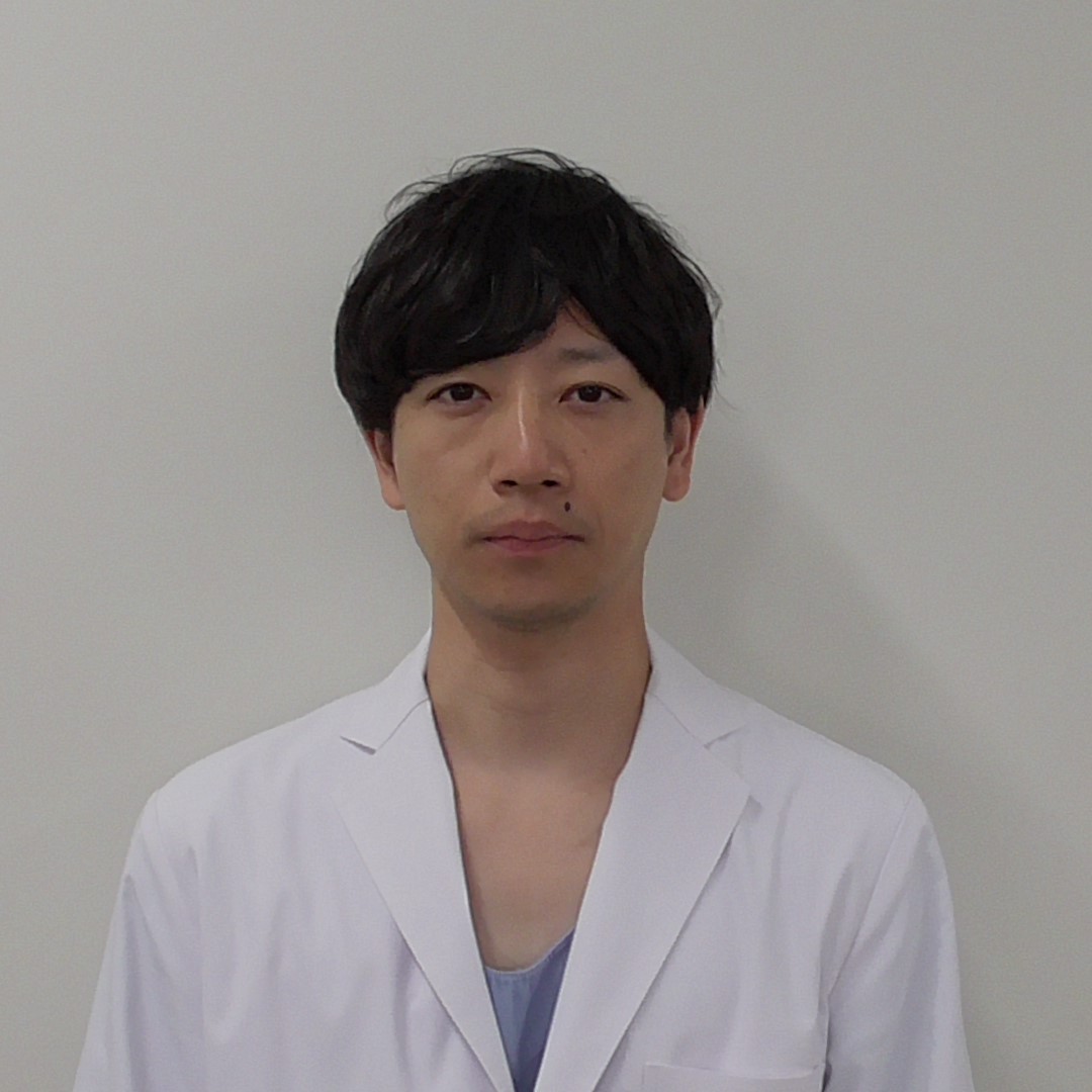 白衣を着た男性が無表情でカメラを見つめているポートレート。背景は無地の壁。