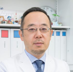 医療白衣を着た男性がカメラを見つめているポートレート。背景には書類や小物が並ぶ棚がある。