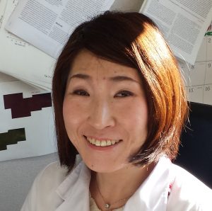 医療現場で微笑む前島裕子教授のポートレート。背景には研究資料が見える。
