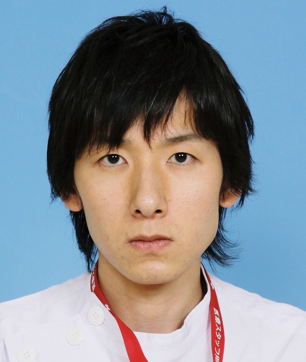 医療従事者の男性が白衣を着て、青い背景の前で真剣な表情をしている写真。