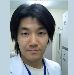 医療用白衣を着た男性の自撮り写真。背景には医療機器が見える。