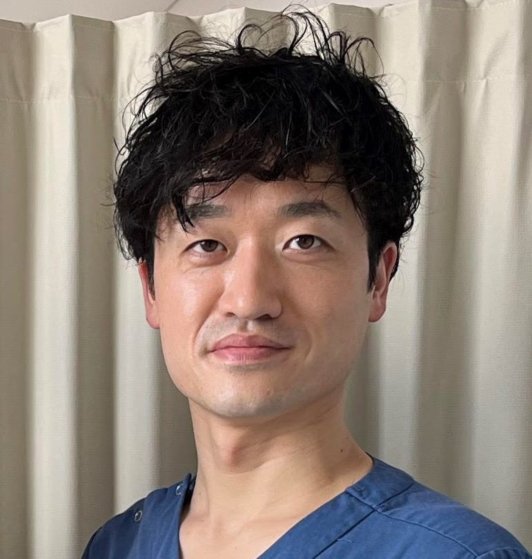 医療現場で働く男性医師が青い医療用衣服を着て、白いカーテンの前で微笑んでいる。