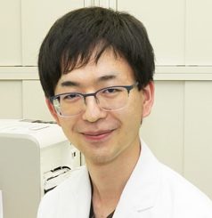 医学部の助手、松本聖生氏が微笑むポートレート。背景には研究機器が見える。
