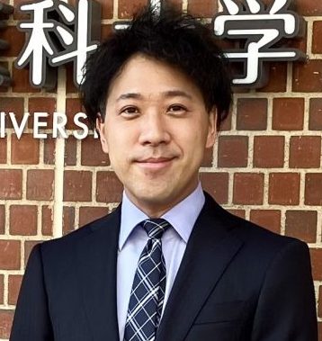 福島県立医科大学の看板の前でスーツを着た男性が微笑んでいる写真。