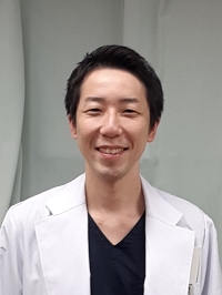 医療白衣を着た男性が微笑んでいるポートレート。背景は無地の壁。