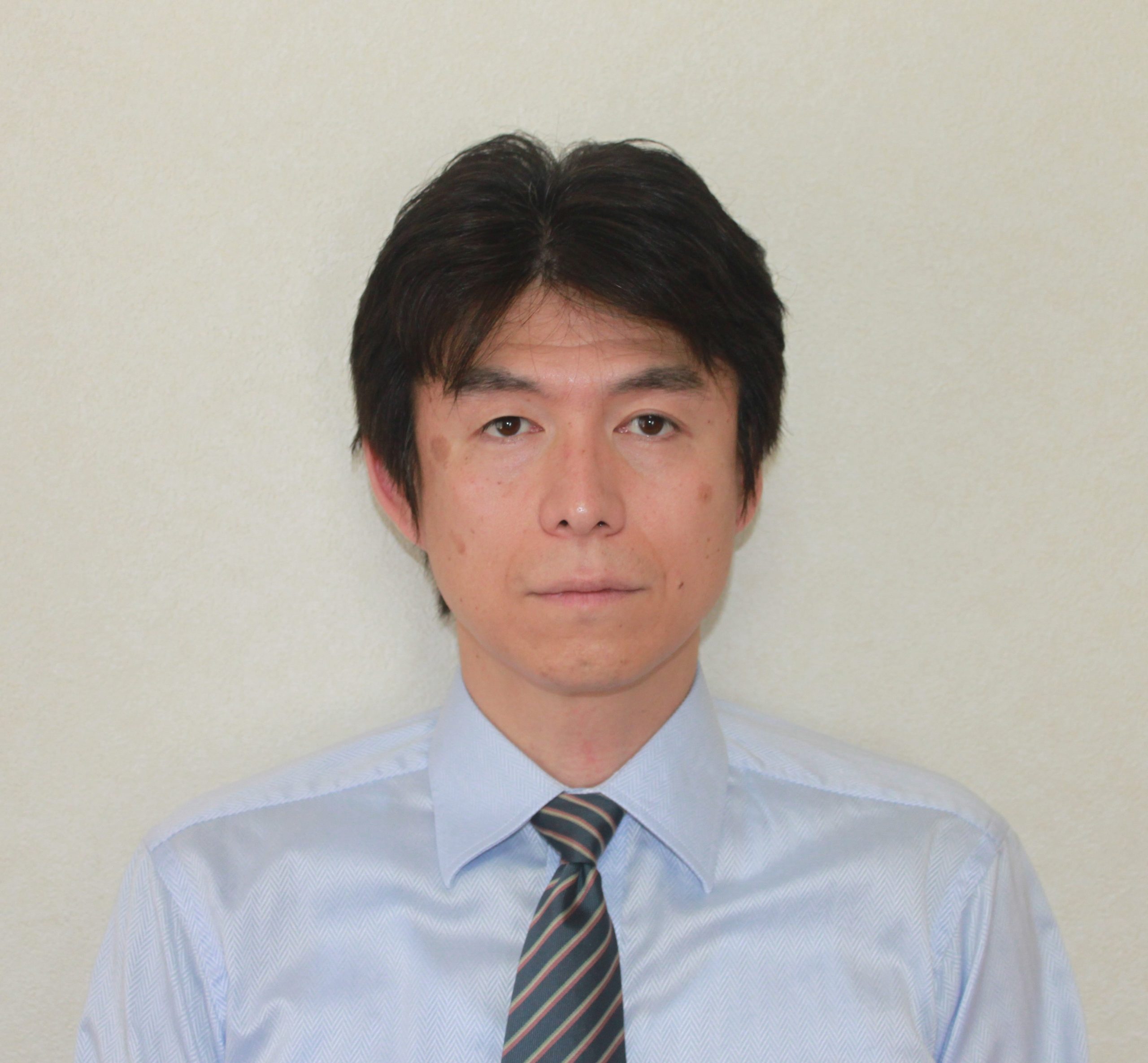 医療に従事する男性がスーツを着て微笑んでいるポートレート。背景は無地の明るい色。
