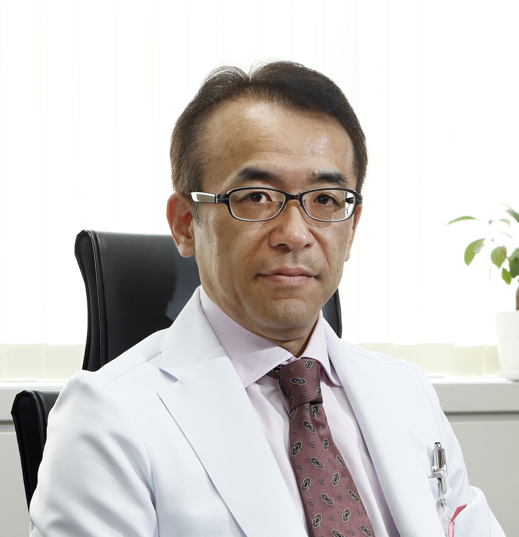 医療白衣を着た男性が椅子に座り、真剣な表情でカメラを見つめている。背景には観葉植物が置かれた明るい室内の様子が見える。
