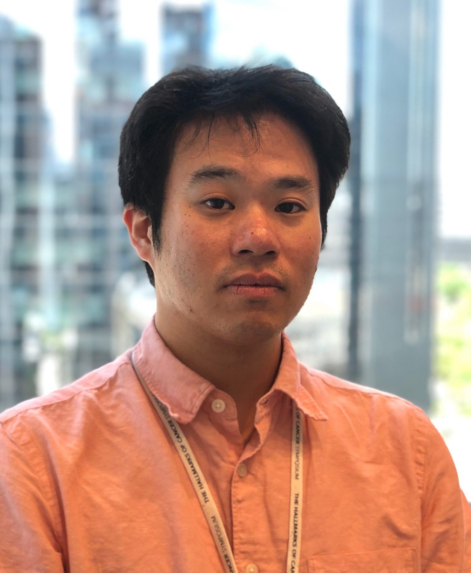 小林信氏がオフィスでカメラに向かって微笑む姿。背景には都市のビル群が見える。