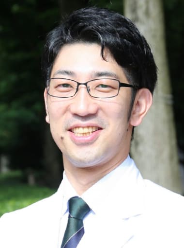 医療白衣を着た男性が笑顔でカメラを見つめている写真。背景には緑の木々が見える。