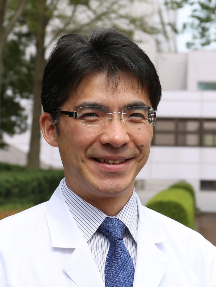 福島県立医科大学の丸橋繁教授が白衣を着て微笑んでいるポートレート。背景には大学の建物が見える。