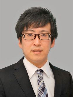 成人発症Still病に関する研究を行う吉田周平助手のポートレート。ビジネススーツを着用し、真剣な表情でカメラを見つめている。
