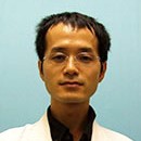 福島県立医科大学の助教、横川哲朗氏のポートレート。白衣を着用し、青い背景の前で微笑んでいる。