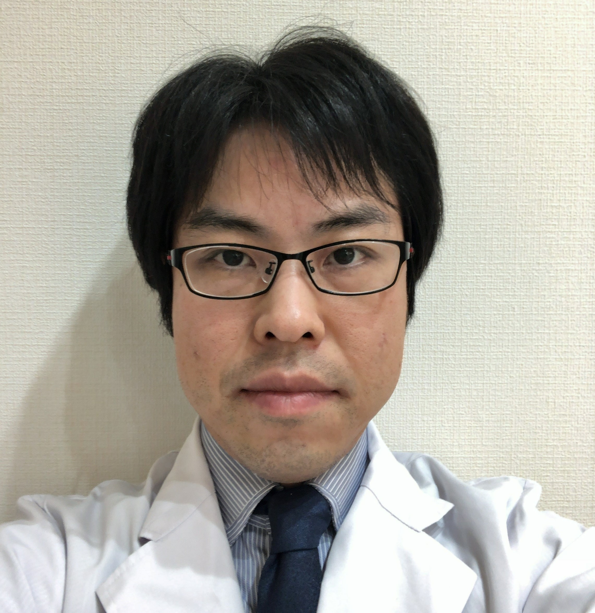 医療用白衣を着た男性がカメラを見つめている自撮り写真。背景は無地の壁。
