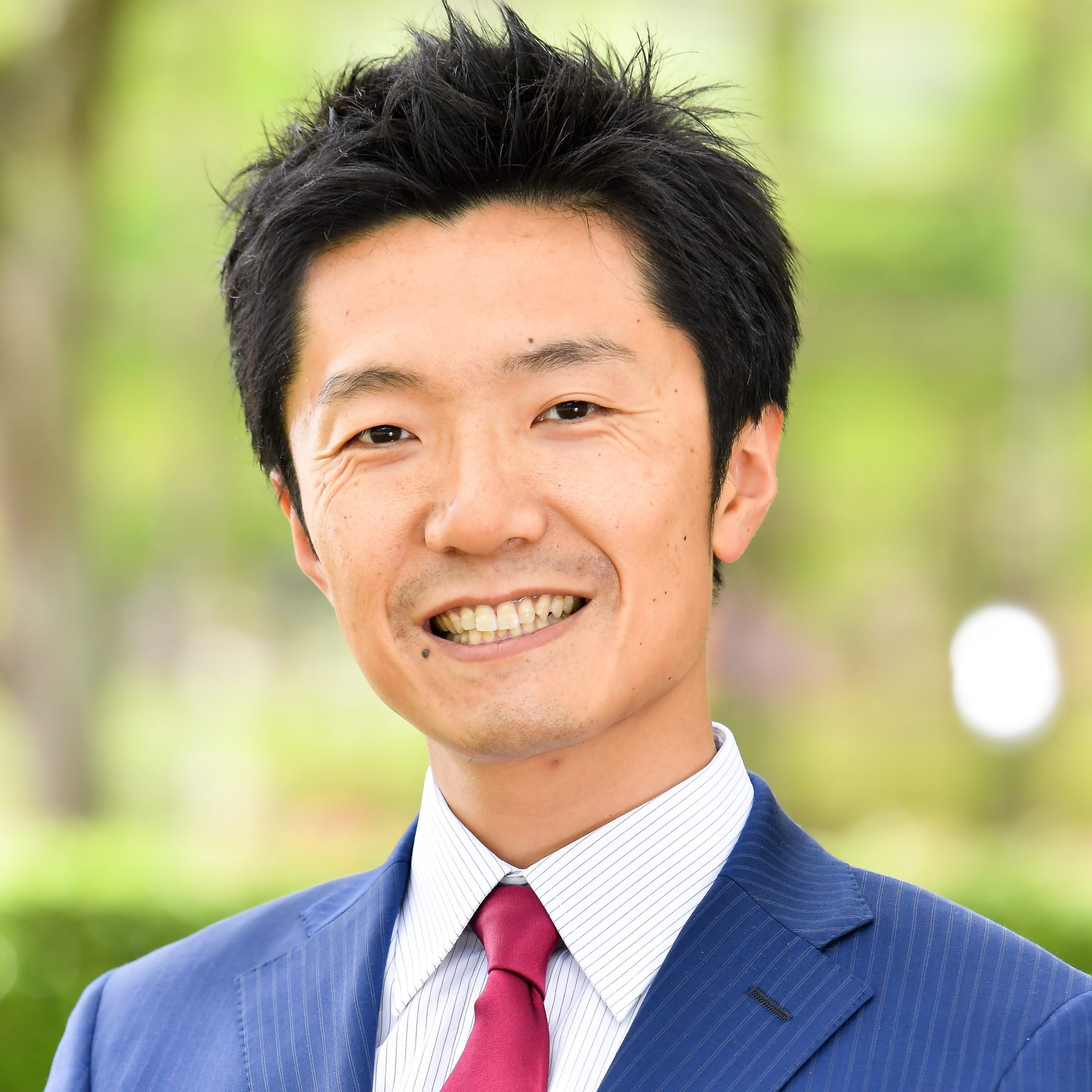 医療に従事する男性が笑顔でポーズをとっている写真。背景には緑の木々が見える。