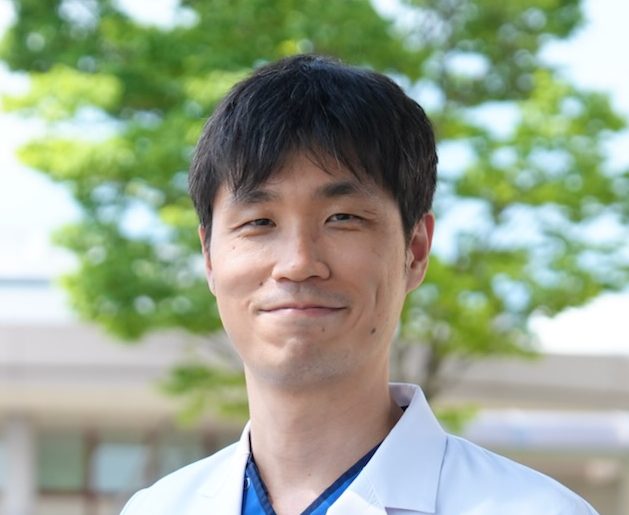医療白衣を着た男性が微笑んでいる写真。背景には緑の木々が見える。