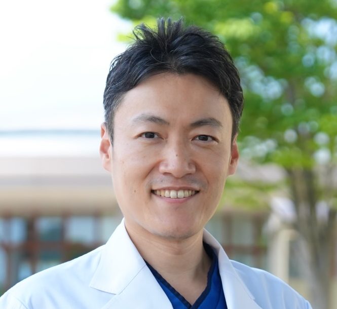 医療白衣を着た男性が笑顔でカメラを見つめている。背景には緑の木々が見える。
