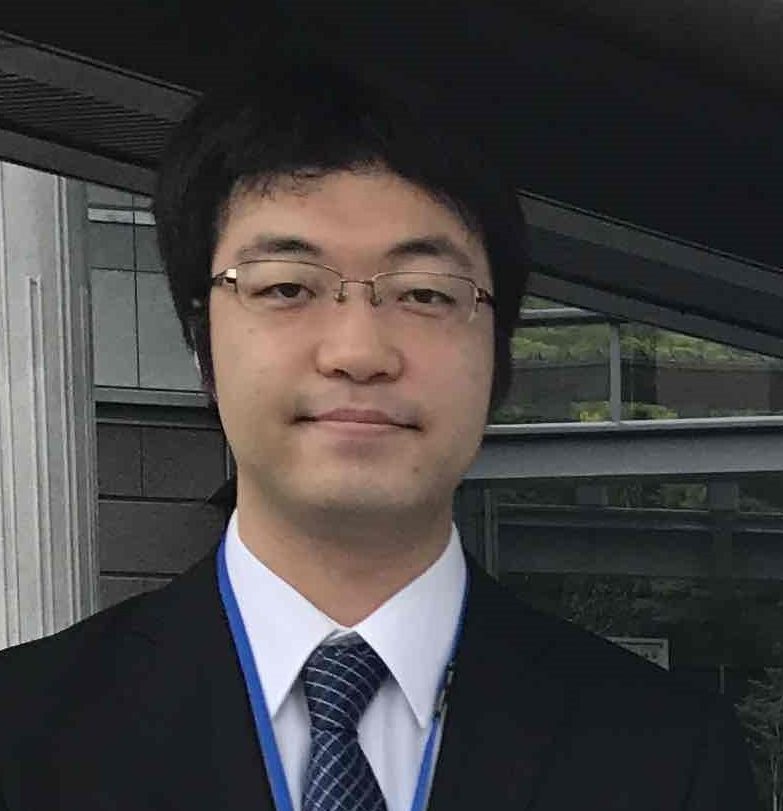 田辺隼人助教がスーツを着て微笑んでいる写真。背景には建物の一部が見える。