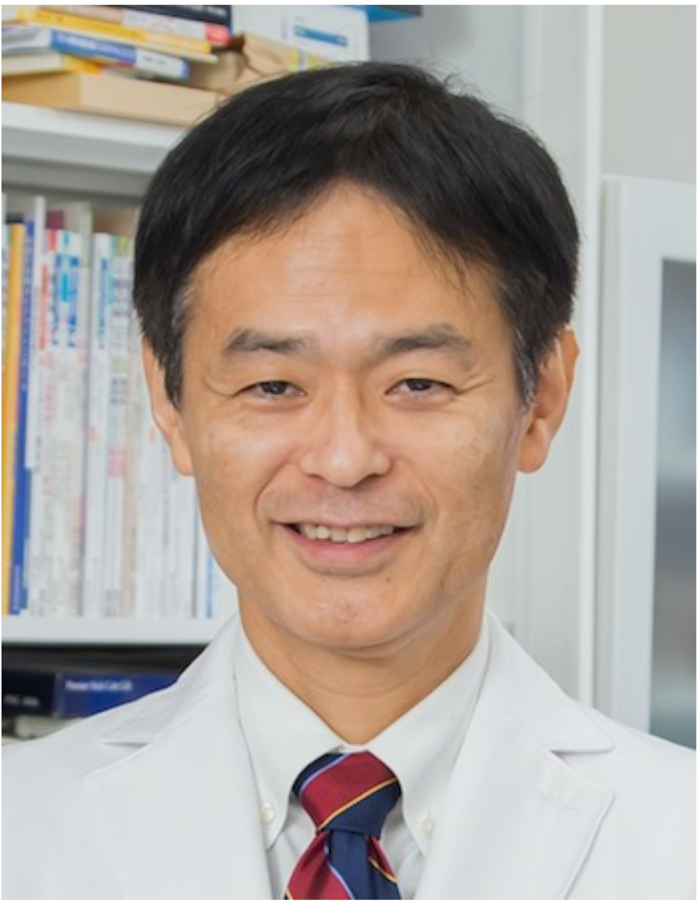 医療白衣を着た男性が微笑んでいるポートレート。背景には書籍が並ぶ棚がある。