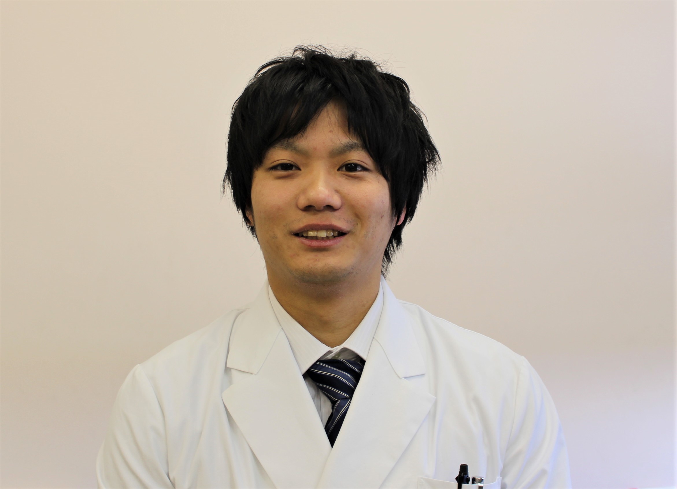白衣を着た男性が微笑んでいるポートレート。背景は淡い色合いでシンプル。