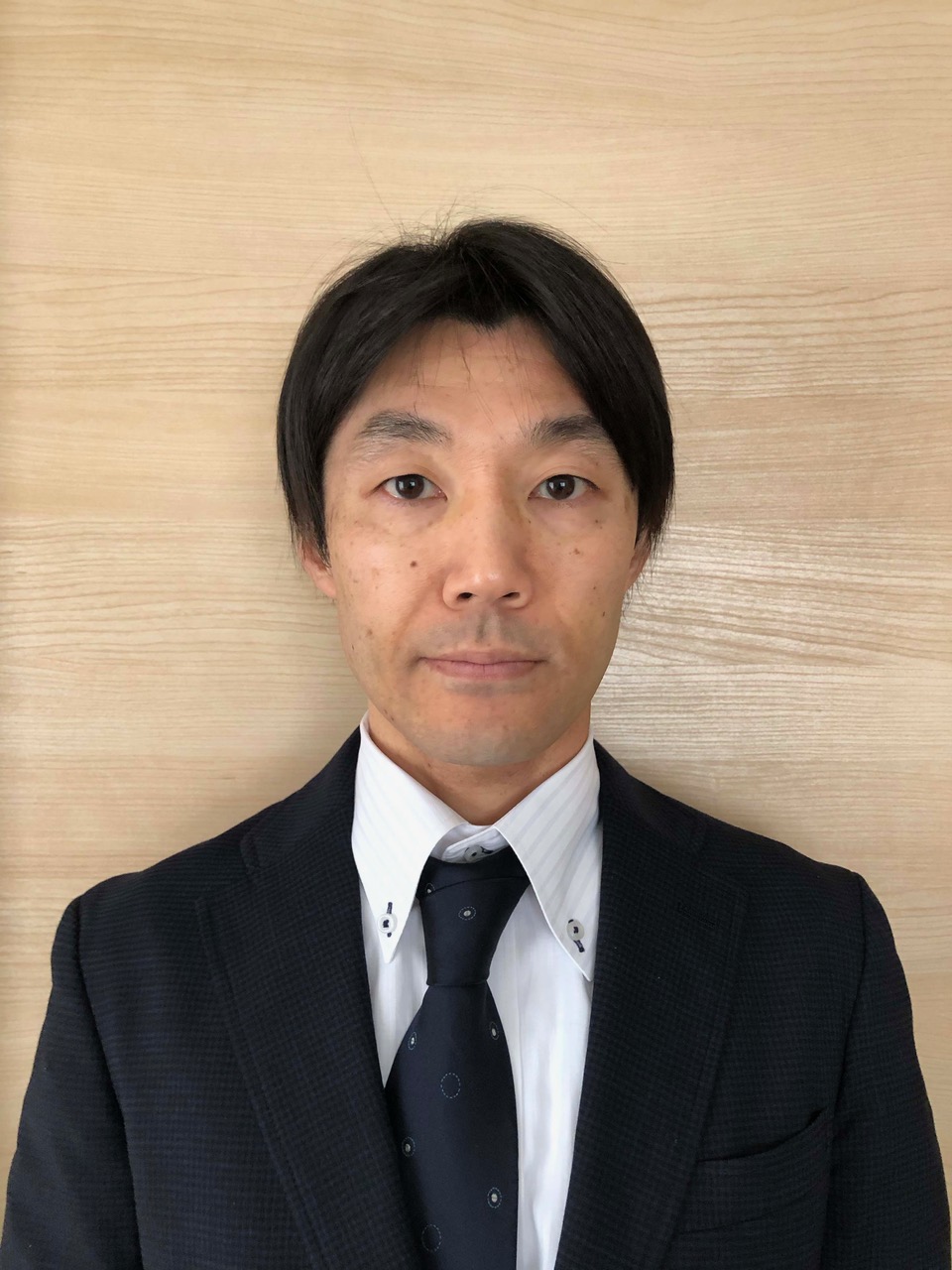 加藤成樹准教授がスーツを着て微笑む姿。背景は木目調の壁。
