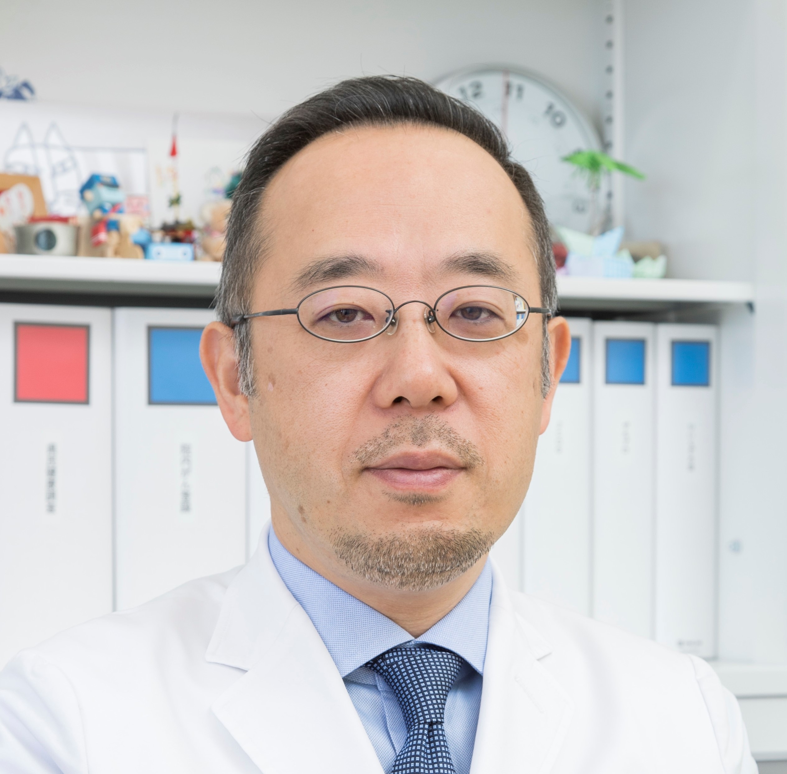 福島県立医科大学の腫瘍内科学講座主任教授、佐治重衡氏のポートレート。白衣を着用し、背景には書類や小物が並ぶ。